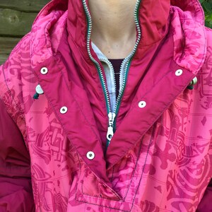 May include: A pink and green patterned jacket with a zipper closure and snap buttons. The jacket has a hood and a high collar.