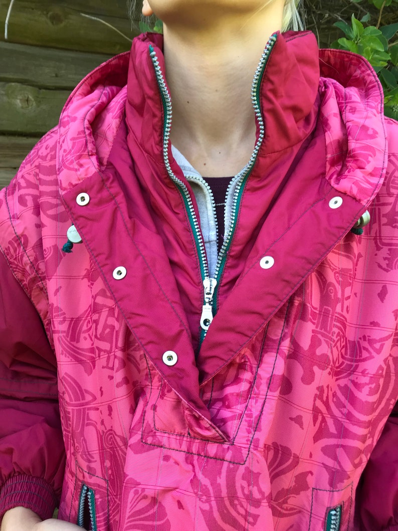 May include: A pink and green patterned jacket with a zipper closure and snap buttons. The jacket has a hood and a high collar.