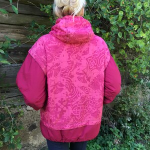 May include: A pink quilted jacket with a patterned design on the back. The jacket has a hood and a zipper closure.