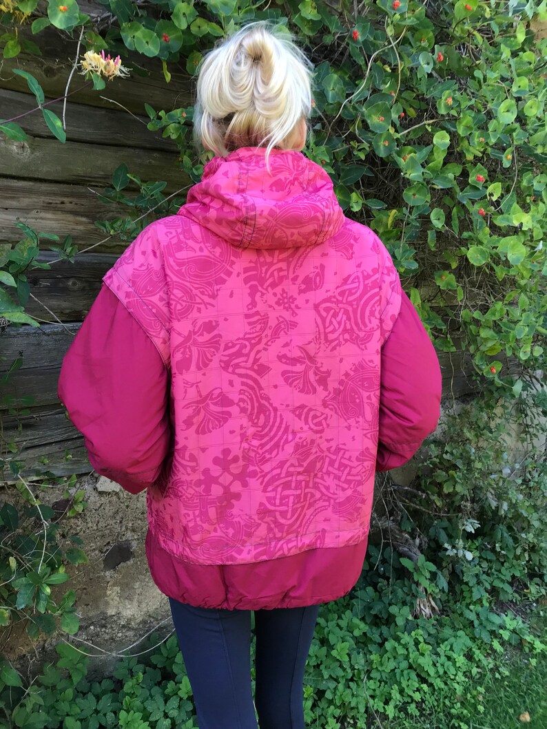 May include: A pink quilted jacket with a patterned design on the back. The jacket has a hood and a zipper closure.