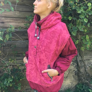 May include: A pink and red patterned jacket with a high collar and a zipper closure. The jacket has a large pocket on the front and a smaller pocket on the sleeve.