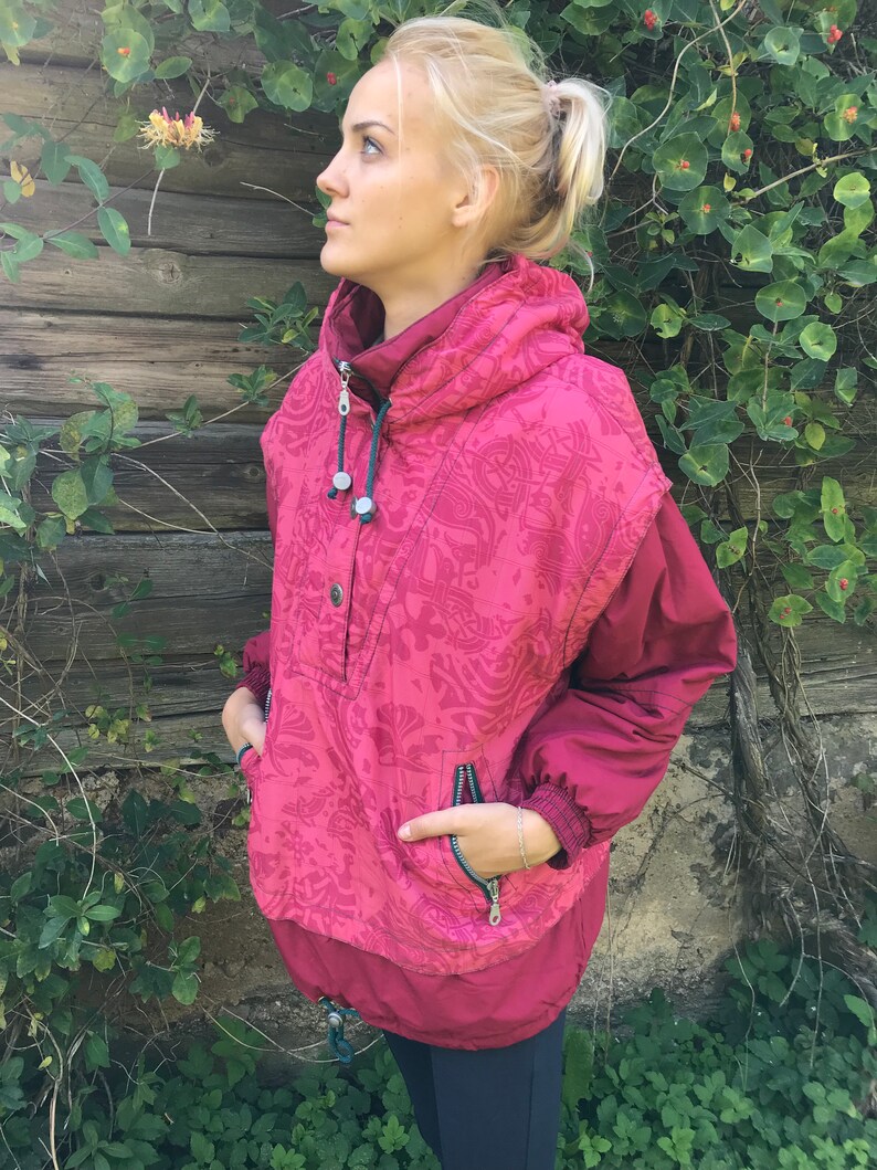 May include: A pink and red patterned jacket with a high collar and a zipper closure. The jacket has a large pocket on the front and a smaller pocket on the sleeve.