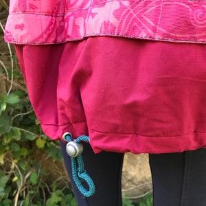 May include: A red jacket with a drawstring at the bottom. The drawstring is teal and has a silver toggle. The jacket is made of a water-resistant material.