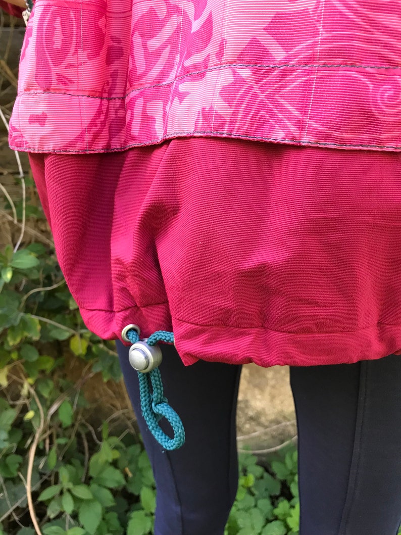 May include: A red jacket with a drawstring at the bottom. The drawstring is teal and has a silver toggle. The jacket is made of a water-resistant material.