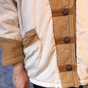 May include: A cream-colored jacket with brown suede accents and brown leather buttons. The jacket has a relaxed fit and a long, flowing silhouette.