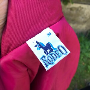 May include: A red jacket with a white label that says "Rodeo" in blue lettering and a donkey illustration. The label also has the number "38" printed on it.