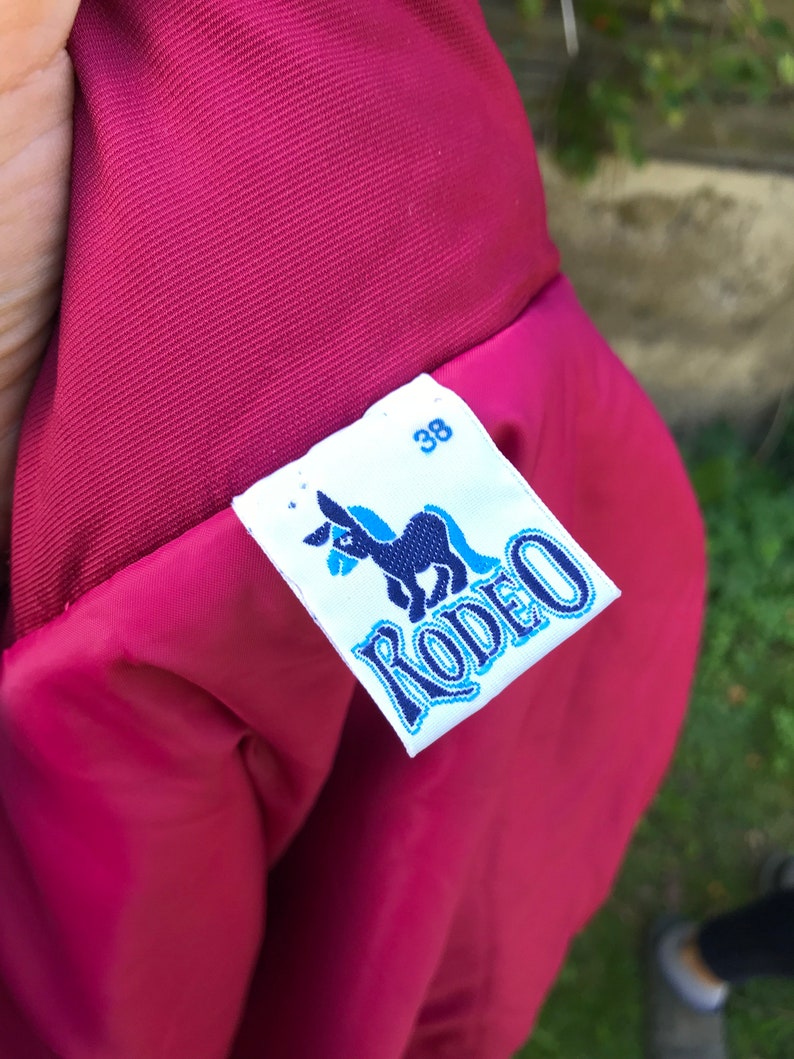 May include: A red jacket with a white label that says "Rodeo" in blue lettering and a donkey illustration. The label also has the number "38" printed on it.