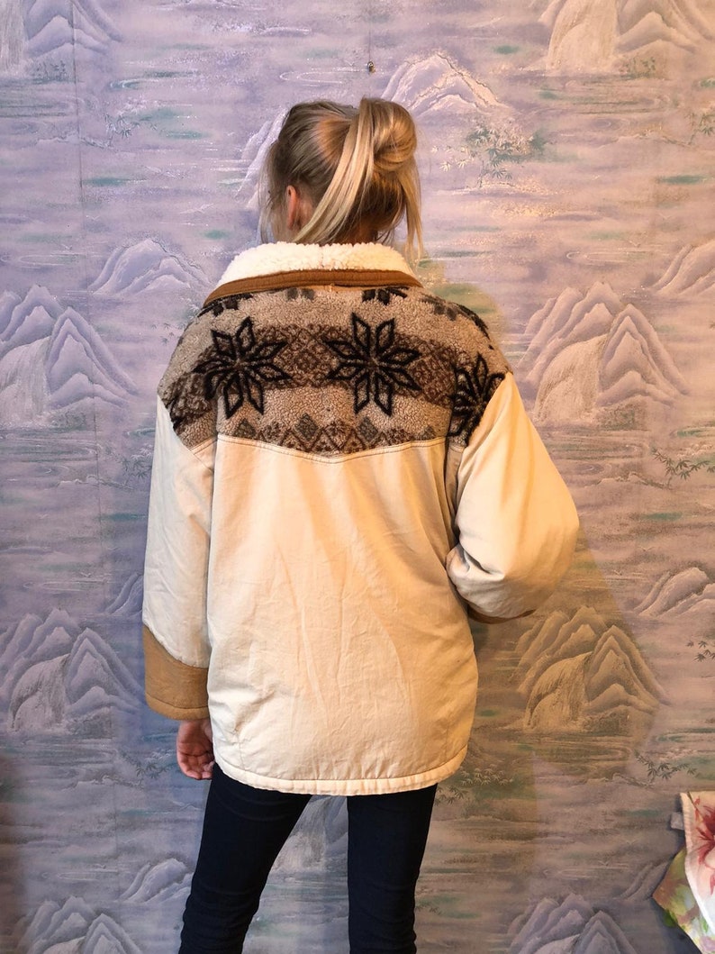 May include: A cream-colored jacket with a brown faux fur collar and a brown and white geometric pattern on the back. The jacket has a zipper closure and long sleeves.