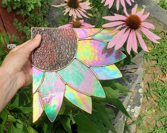 May include: A stained glass sunflower with iridescent colors, featuring a dark brown center and pink, green, and blue petals. The sunflower is displayed against a backdrop of green foliage and pink flowers.