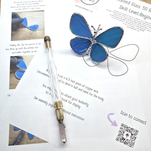 May include: A stained glass butterfly with blue wings and a silver metal body, positioned on a printed instructional guide titled Stained Glass 3D Butterfly. A glass cutter tool with a clear handle and metal tip rests diagonally across the paper.