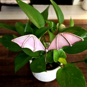 May include: A decorative plant stake featuring pink stained glass bat wings with copper-colored edging. The wings are set in a lush green plant in a white pot, creating a whimsical garden accent. The stake is designed to add a touch of fantasy to indoor or outdoor spaces.