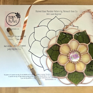May include: A stained glass mandala suncatcher with green and cream petals, a central iridescent cabochon, and copper foil solder lines. It includes a copper chain, a glass cutter tool, and a printed pattern sheet titled Stained Glass Mandala Pattern by Tetreault Glass Co. Skill Level: Beginner.