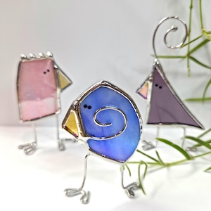 May include: Three colorful stained glass bird sculptures. The birds have silver wire legs and beaks. One bird is pink, one is blue with a swirl, and the other is purple. The birds are on a white surface.