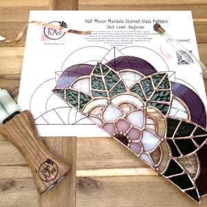 May include: A half-moon stained glass pattern for beginners, with purple, green, white, and pink glass pieces. A wooden-handled tool and a glass cutter are also present. The pattern is on a white sheet of paper.