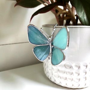 May include: A stained glass butterfly with a silver-colored metal frame. The butterfly's wings are a gradient of blue and teal. The butterfly is hanging near a white pot with a plant.