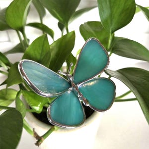 May include: A teal-colored butterfly decoration with a silver-colored border. The butterfly has four wings and is set against a background of green leaves and a white pot. The butterfly is a decorative item.