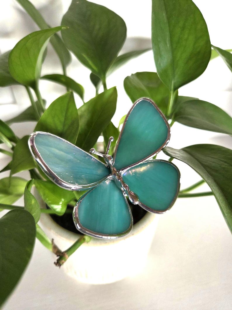 May include: A teal-colored butterfly decoration with a silver-colored border. The butterfly has four wings and is set against a background of green leaves and a white pot. The butterfly is a decorative item.