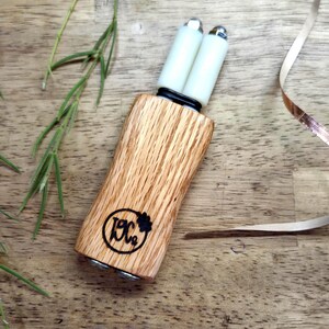 May include: A handheld tool featuring a light brown wood handle with a visible grain pattern and a black circular logo stamped on the front. Two white cylindrical rollers with metallic silver caps are attached to the top, secured by a black band.