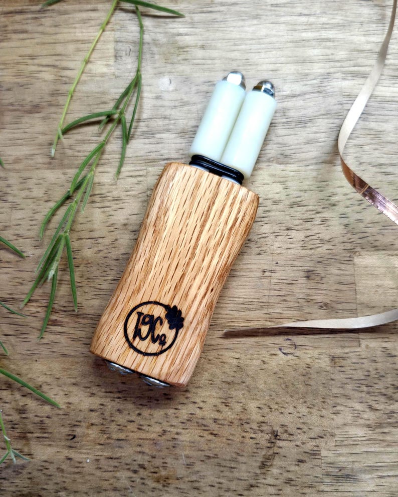 May include: A handheld tool featuring a light brown wood handle with a visible grain pattern and a black circular logo stamped on the front. Two white cylindrical rollers with metallic silver caps are attached to the top, secured by a black band.