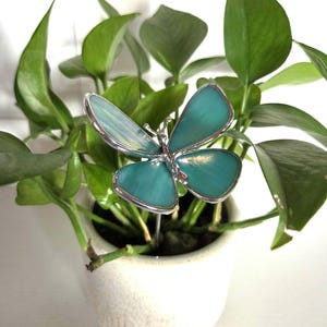May include: A teal and silver butterfly plant stake is inserted into a small white ceramic pot filled with green leafy plants. The butterfly has a detailed design with a silver outline and a teal-colored body. The plant pot is round and has a textured surface.