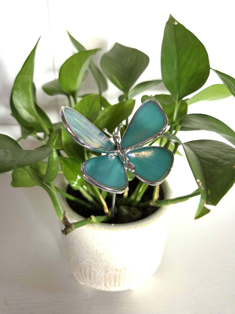 May include: A teal and silver butterfly plant stake is inserted into a small white ceramic pot filled with green leafy plants. The butterfly has a detailed design with a silver outline and a teal-colored body. The plant pot is round and has a textured surface.