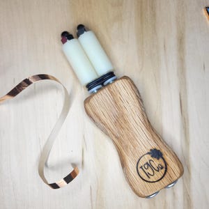 May include: A handheld tool with a contoured wooden handle featuring a circular logo with the text T9Co. Two white cylindrical rollers are attached to the top, secured by black rubber rings and metal hardware. A thin strip of copper foil lies nearby.