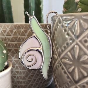 May include: A stained glass snail plant stake with a silver metal border. The snail's shell is a swirl of white and pink, and its body is light green. The stake is attached to a gray ceramic pot with a diamond pattern, with a cactus in the background.