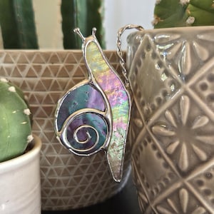 May include: A stained glass snail suncatcher with iridescent shell and silver solder. The snail is hanging near a gray ceramic pot with a diamond pattern. The background includes green cacti in white and gray pots.
