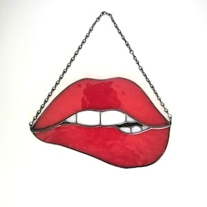 May include: A stained glass piece depicting a pair of red lips with white teeth and a black bite mark. The artwork is suspended by a black chain against a white background. The lips have a glossy finish.