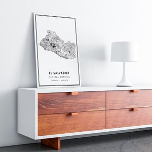 May include: A black and white map print of El Salvador, Central America, with the coordinates 13.7942&deg;N 88.8963&deg;W. The print is framed and hanging on a white wall above a white dresser with brown drawers.