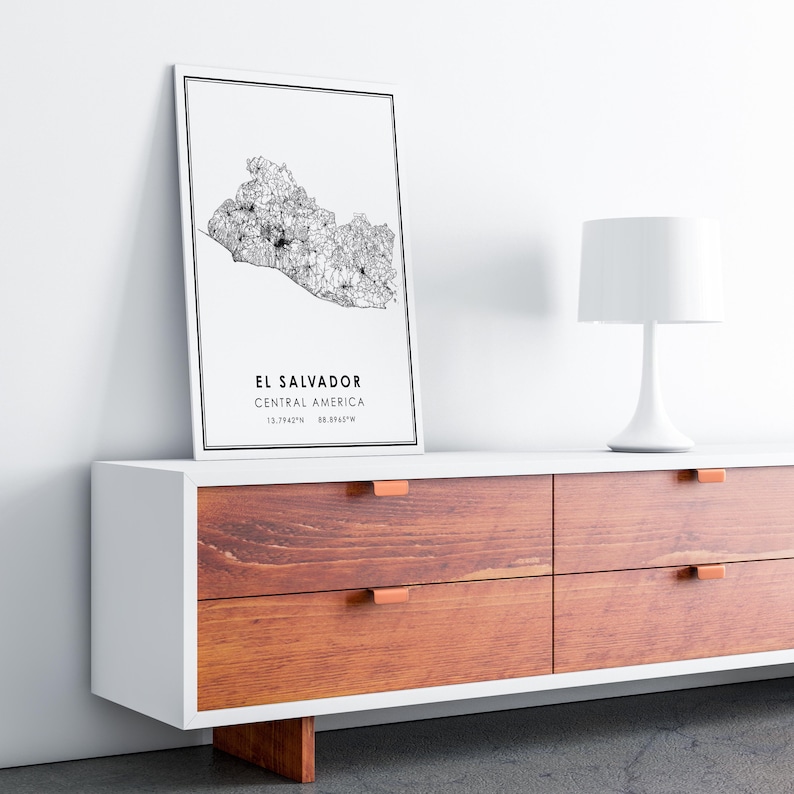 May include: A black and white map print of El Salvador, Central America, with the coordinates 13.7942&deg;N 88.8963&deg;W. The print is framed and hanging on a white wall above a white dresser with brown drawers.