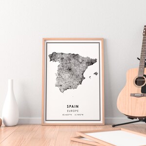 May include: A framed black and white map print of Spain with the text "SPAIN EUROPE 40.4637&deg;N 3.7492&deg;W" printed below the map. The print is hanging on a white wall next to a wooden guitar stand with a brown acoustic guitar.