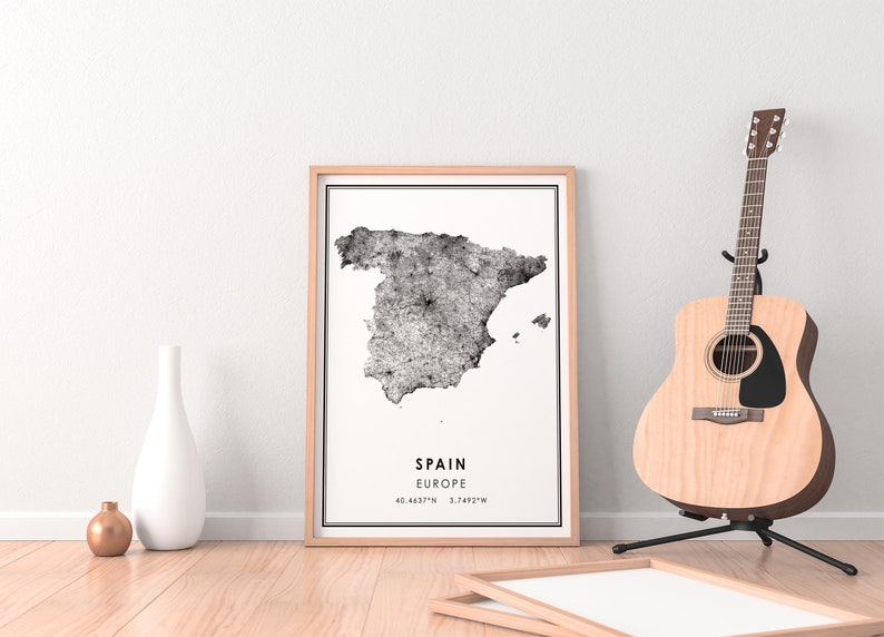 May include: A framed black and white map print of Spain with the text "SPAIN EUROPE 40.4637&deg;N 3.7492&deg;W" printed below the map. The print is hanging on a white wall next to a wooden guitar stand with a brown acoustic guitar.