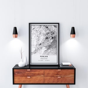 May include: Black and white map print of Punjab, Pakistan, with the coordinates 31.1764&deg;N 72.0597&deg;E. The print is framed in a black frame and hanging on a white wall above a wooden dresser with black drawer fronts.