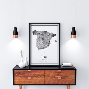 May include: Black and white print of a map of Spain with the text "SPAIN EUROPE 40.4637&deg;N 3.7492&deg;W" on a white background. The print is framed in black and hanging on a white wall above a wooden dresser with black drawer fronts.