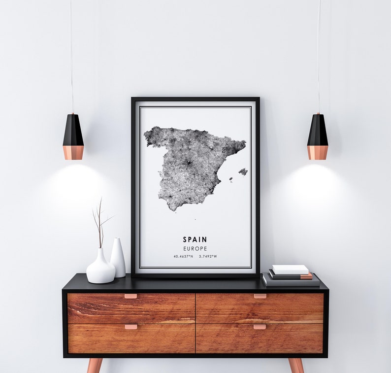 May include: Black and white print of a map of Spain with the text "SPAIN EUROPE 40.4637&deg;N 3.7492&deg;W" on a white background. The print is framed in black and hanging on a white wall above a wooden dresser with black drawer fronts.
