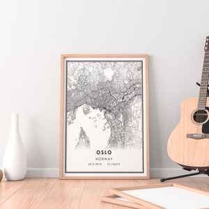 May include: A black and white map print of Oslo, Norway in a light wood frame. The map shows the city streets and waterways. The print is hanging on a white wall with a wooden floor and a guitar in the background.