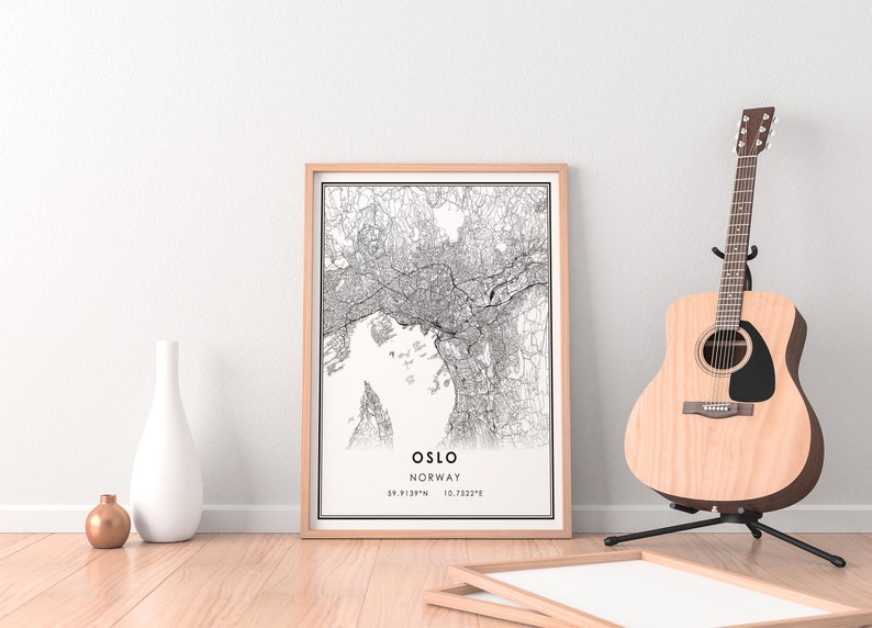May include: A black and white map print of Oslo, Norway in a light wood frame. The map shows the city streets and waterways. The print is hanging on a white wall with a wooden floor and a guitar in the background.