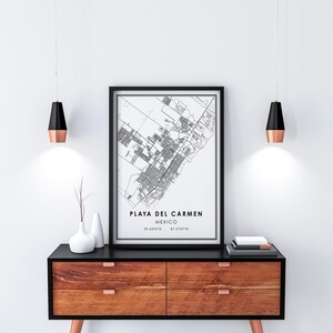May include: A framed black and white map print of Playa del Carmen, Mexico, displayed on a wooden cabinet. The print includes the city's name and coordinates. Two modern pendant lights with black and copper finishes hang above the cabinet.