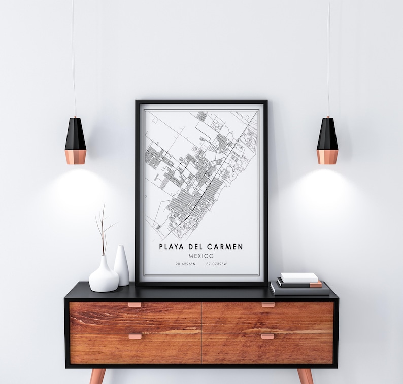 May include: A framed black and white map print of Playa del Carmen, Mexico, displayed on a wooden cabinet. The print includes the city's name and coordinates. Two modern pendant lights with black and copper finishes hang above the cabinet.