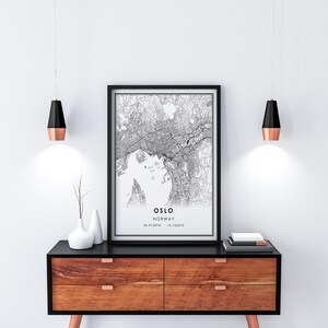 May include: A black and white map print of Oslo, Norway, with the city's name and coordinates printed below the map. The print is framed in a black frame and is hanging on a white wall above a wooden dresser with black drawer fronts.