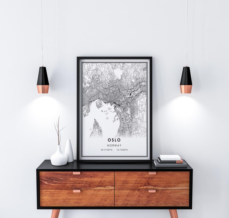 May include: A black and white map print of Oslo, Norway, with the city's name and coordinates printed below the map. The print is framed in a black frame and is hanging on a white wall above a wooden dresser with black drawer fronts.
