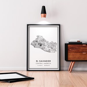 May include: A black and white line art map print of El Salvador, Central America. The print is framed in a black frame and hangs on a white wall. The coordinates 13.7942&deg;N 88.8965&deg;W are printed below the map.