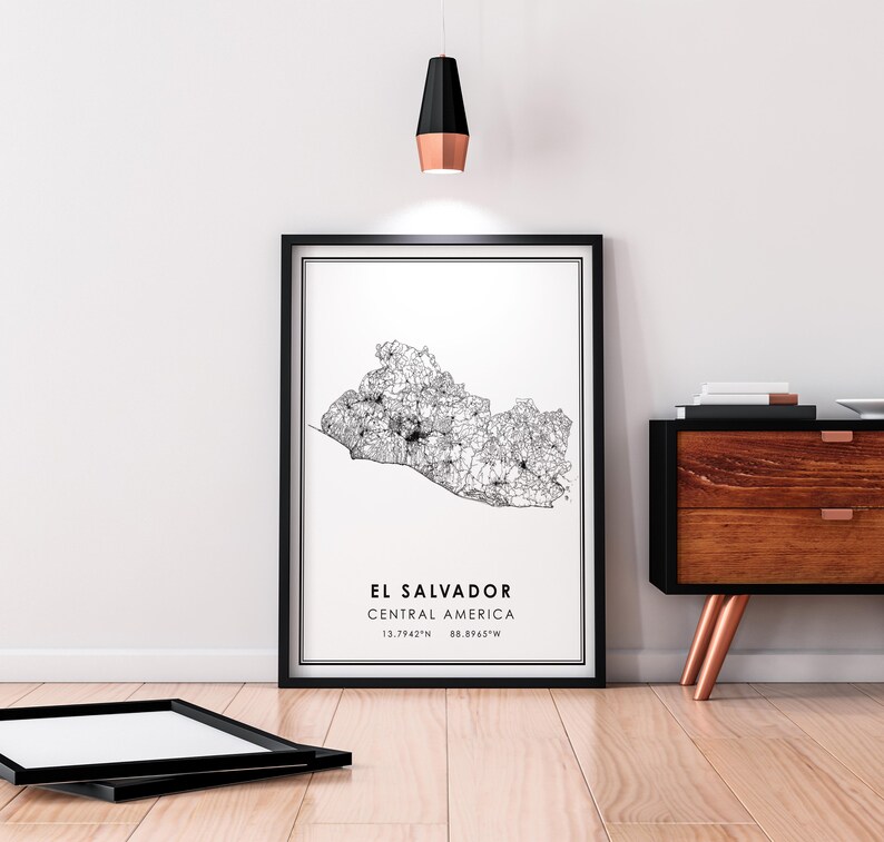 May include: A black and white line art map print of El Salvador, Central America. The print is framed in a black frame and hangs on a white wall. The coordinates 13.7942&deg;N 88.8965&deg;W are printed below the map.