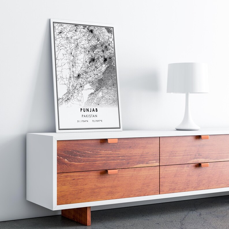May include: A black and white map print of Punjab, Pakistan, with the text "PUNJAB PAKISTAN 31.1364&deg;N 72.7097&deg;E" at the bottom. The print is framed and hanging on a white wall above a white dresser with four drawers. The dresser has a wood grain finish and copper-colored drawer pulls.