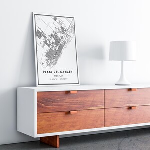 May include: A framed black and white map print of Playa del Carmen, Mexico, displayed on a white and wood-toned cabinet. The print includes the city's name and coordinates. A white table lamp sits to the right.