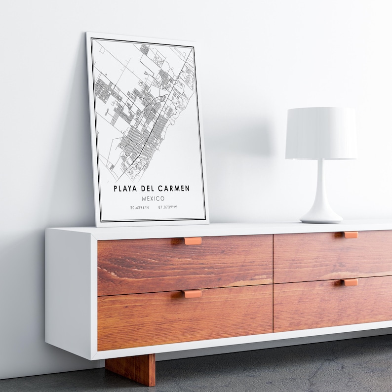 May include: A framed black and white map print of Playa del Carmen, Mexico, displayed on a white and wood-toned cabinet. The print includes the city's name and coordinates. A white table lamp sits to the right.