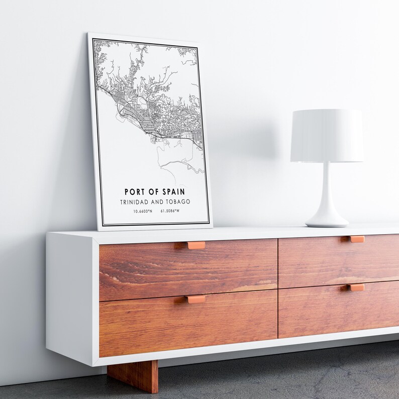 Puede incluir: Una impresi&oacute;n de mapa en blanco y negro de Puerto Espa&ntilde;a, Trinidad y Tobago, con las coordenadas de la ciudad enumeradas debajo. La impresi&oacute;n est&aacute; enmarcada y colgada en una pared blanca sobre una c&oacute;moda de madera con tiradores de cajones de cobre.