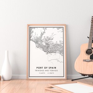 Puede incluir: Una impresi&oacute;n de mapa en blanco y negro de Puerto Espa&ntilde;a, Trinidad y Tobago, enmarcada en un marco de madera clara. El mapa est&aacute; sobre un fondo blanco e incluye las coordenadas 10.6465&deg;N 61.5084&deg;W. La impresi&oacute;n est&aacute; sobre un suelo de madera con un jarr&oacute;n blanco y un jarr&oacute;n marr&oacute;n delante de ella. Una guitarra ac&uacute;stica marr&oacute;n est&aacute; en el fondo.