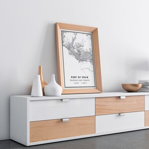 Puede incluir: Un mapa en blanco y negro enmarcado de Puerto Espa&ntilde;a, Trinidad y Tobago, con las coordenadas 10.6489&deg;N, 61.5088&deg;W impresas debajo del mapa. El marco es de madera clara y el mapa est&aacute; sobre una c&oacute;moda blanca de seis cajones.